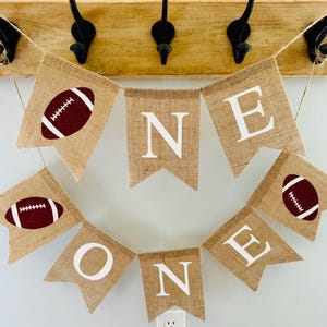 May include: A burlap banner with the word "ONE" spelled out in white letters. The banner is decorated with brown footballs and is strung on a twine.
