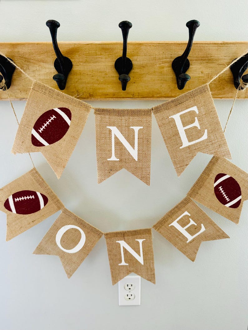 May include: A burlap banner with the word "ONE" spelled out in white letters. The banner is decorated with brown footballs and is strung on a twine.