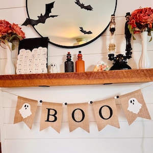 May include: A burlap banner featuring five pennants strung on a cord with black and white beads. The two outer pennants display white ghost illustrations, while the three center pennants feature the black letters B, O, and O.
