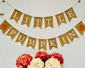 May include: A burlap banner with the words "Little Pumpkin" in white letters. The banner is decorated with small orange pumpkins. The banner is hanging on a white wall above a wooden table with three vases of white and pink hydrangeas.
