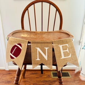May include: A burlap banner with the word "ONE" and a football design in maroon and white. The banner is strung across a wooden high chair.