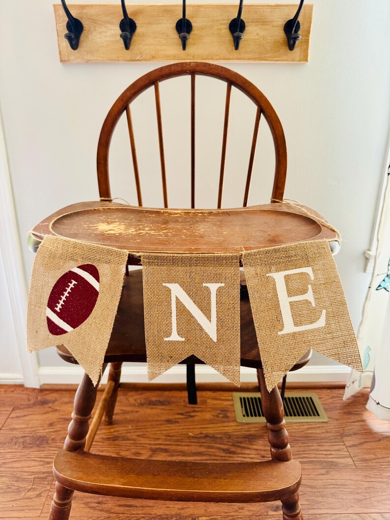 May include: A burlap banner with the word "ONE" and a football design in maroon and white. The banner is strung across a wooden high chair.