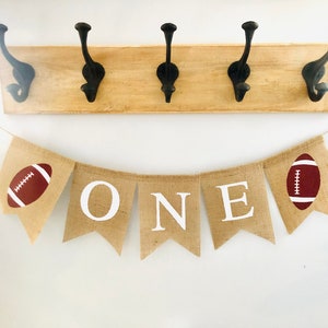 May include: A burlap banner with the word "ONE" in white letters and two brown footballs on either end. The banner is strung on a white wall with a wooden rack with four black hooks above it.