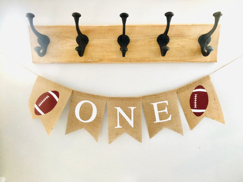 May include: A burlap banner with the word "ONE" in white letters and two brown footballs on either end. The banner is strung on a white wall with a wooden rack with four black hooks above it.