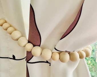 May include: A set of wooden beads strung together to create a curtain tieback. The beads are a light natural wood color and are strung on a white cord. The tieback is holding back a white curtain with a brown and white pattern.