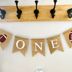 May include: A burlap banner with the word "ONE" in white letters and two brown footballs on either end. The banner is strung up on a white wall.