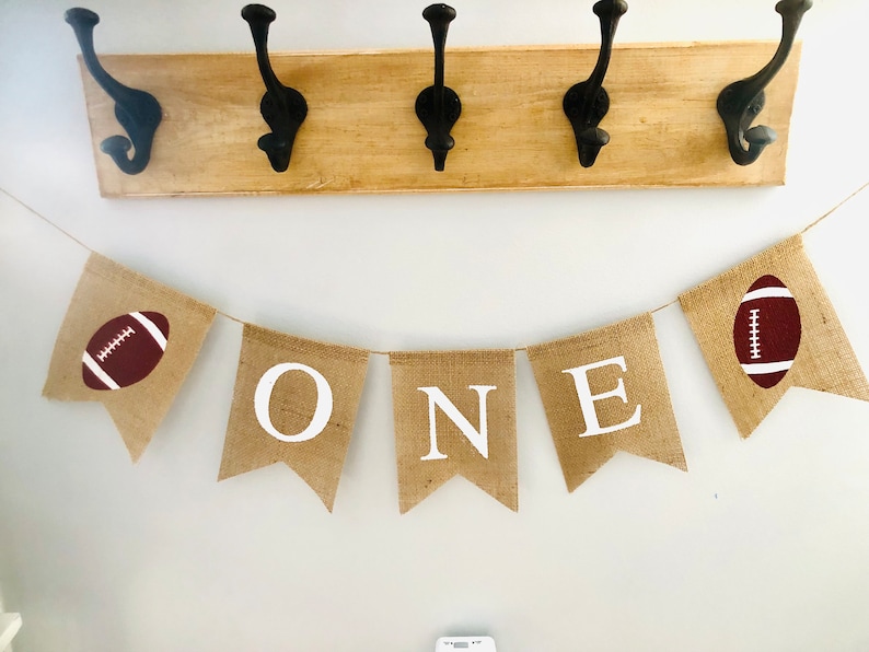 May include: A burlap banner with the word "ONE" in white letters and two brown footballs on either end. The banner is strung up on a white wall.