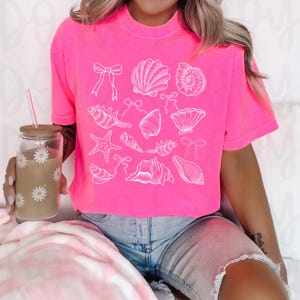 May include: A pink cropped t-shirt with a white graphic of seashells, starfish, and bows.
