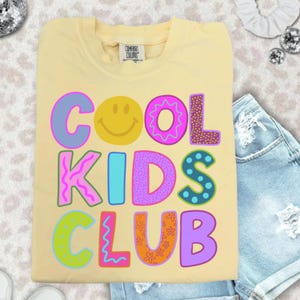 Peut inclure: Un t-shirt jaune clair Comfort Colors avec l'inscription "COOL KIDS CLUB" en lettres colorées et bombées. Le mot "COOL" contient un smiley. Le t-shirt est plié et posé sur une surface claire avec un short en jean.