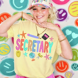 May include: A light yellow t-shirt featuring the word SECRETARY in colorful, patterned block letters. The design includes illustrations of a pencil, a notebook labeled Notes, a globe, scissors, and stars. The shirt is worn with a multi-colored hat.
