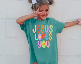 May include: A teal t-shirt with the phrase "Jesus Loves You" in bright, multi-coloured letters. The design includes a smiley face and star details. The child is wearing a white bow and smiling, making a peace sign.