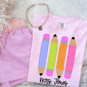 Könnte beinhalten: Ein hellrosa T-Shirt mit einer Grafik von vier bunten Stiften mit Mustern und dem Text "Mrs. Jones" in schwarzer Schrift. Das T-Shirt wird mit einer rosa-weiß karierten Shorts und einer goldfarbenen Kette mit rundem Anhänger kombiniert.
