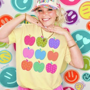 K&ouml;nnte beinhalten: Ein hellgelbes T-Shirt mit einem Raster aus neun stilisierten &Auml;pfeln in verschiedenen Farben wie Pink, Gr&uuml;n, Lila, Blau und Orange. Jeder Apfel hat ein einzigartiges Muster wie Sterne, Punkte oder Blumen und ein einfaches Umrissdesign.