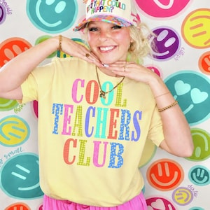 May include: A pale yellow short-sleeve t-shirt featuring the text COOL TEACHERS CLUB printed in large, multi-colored block letters with various patterns including stars and dots. The shirt is paired with a beige trucker hat with colorful text.
