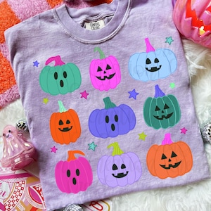 May include: A light purple short-sleeve t-shirt featuring a graphic print of nine colorful jack-o'-lanterns and ghosts with various facial expressions in shades of teal, pink, blue, orange, and purple, accented by small scattered stars.