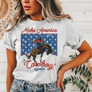 May include: A grey t-shirt with a vintage-style graphic of a cowboy riding a horse in front of an American flag. The text "Make America Cowboy Again" is printed on the shirt.