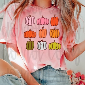 May include: A light pink t-shirt featuring a grid of nine cartoon pumpkins in various colors, including pink, orange, green, and yellow. The pumpkins have black outlines and green stems. The shirt is a casual style.