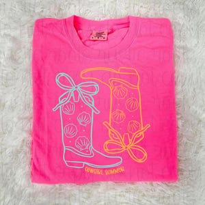 May include: A bright pink t-shirt featuring a graphic of two cowboy boots with seashell designs and bows. The boots are outlined in light blue and yellow, with the words "COWGIRL SUMMER" printed at the bottom.