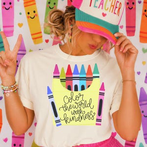 May include: A cream-colored t-shirt featuring a graphic of a yellow crayon box filled with colorful crayons and the text "Color the world with kindness." The shirt is paired with a multi-colored trucker hat with the word "TEACHER" printed on the front.