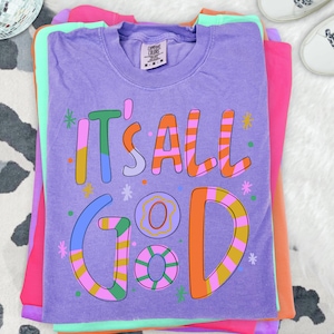 Può includere: Una pila di magliette colorate. La maglietta superiore è viola con la frase "IT'S ALL GOOD" in un carattere giocoso e multicolore. Altre magliette nella pila sono rosa, arancioni e verdi. Le magliette sono su una superficie con stampa di mucca bianca e nera.