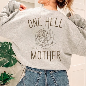 May include: A light gray sweatshirt with the phrase "ONE HELL OF A MOTHER" printed on the back in brown lettering. A detailed rose illustration is centered below the text. The sweatshirt is made of a soft material.