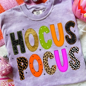 May include: A light purple t-shirt with the text HOCUS POCUS printed in large, multi-colored letters. The letters feature various patterns including spiderwebs, gold glitter, green polka dots, solid orange, candy corn, leopard print, and ghosts.