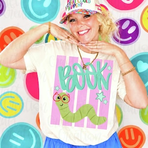 May include: A cream-colored t-shirt featuring a graphic of a green worm wearing red glasses. Above the worm, the word BOOK is written in large, teal, hand-lettered font. The design is set against a background of vertical pink stripes and small teal stars.