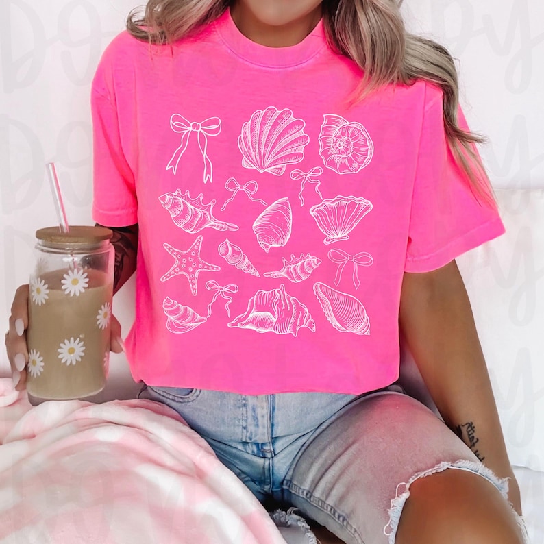 May include: A pink cropped t-shirt with a white graphic of seashells, starfish, and bows.