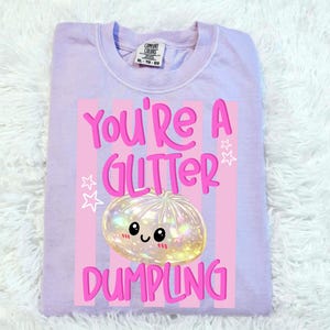May include: A light purple Comfort Colors XL-TG-EG sweatshirt. The front features a graphic with the text "You're a Glitter Dumpling." The graphic includes a cute, glittery dumpling with a smiling face, set against pink stripes and stars.