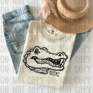 May include: Cream-colored t-shirt featuring a black and white alligator graphic with the word "Gators" below. The shirt is paired with light-wash denim jeans and a tan hat. A necklace is visible on the jeans.