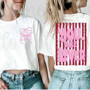 Könnte beinhalten: Weißes Crop-Top mit einer rosa Schleife und dem Text "JOHN 3:16" auf der Vorderseite. Auf der Rückseite des Shirts stehen die Worte "YOU ARE LOVED" in Rosa auf einem rot-weiß gestreiften Hintergrund.