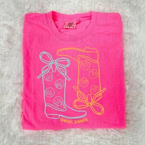 May include: A bright pink t-shirt featuring a graphic of two cowboy boots with seashell designs and bows. The boots are outlined in light blue and yellow, with the words "COWGIRL SUMMER" printed at the bottom.