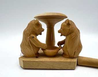 Peut inclure: Un jouet mécanique en bois brun clair représentant deux ours sculptés face à face avec un bol central et un sommet en forme de champignon. Les ours sont montés sur une base plate avec une poignée en bois sur le côté pour le mécanisme.