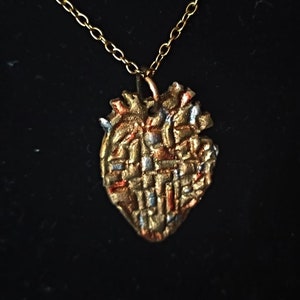 Könnte beinhalten: Eine goldene Halskette mit einem herzförmigen Anhänger mit strukturierter Oberfläche. Der Anhänger ist aus Metall und hat eine goldene, silberne und kupferne Oberfläche.