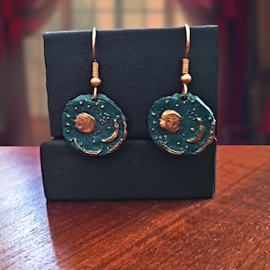 Op de afbeelding: Een paar oorbellen met ronde, blauwgroene schijfjes met een getextureerd oppervlak. Elke schijf is versierd met een verhoogd, goudkleurig motief van een zon en een maansikkel. De oorbellen hangen aan koperkleurige metalen haakjes.