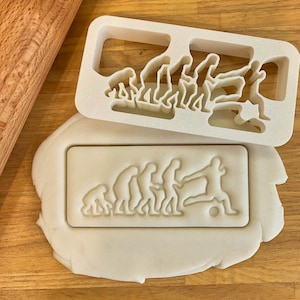 Könnte beinhalten: Ein weißer Plastik-Ausstecher mit einem Design aus einer Reihe von Strichmännchen, die Fußball spielen. Der Ausstecher liegt auf einem weißen, ausgerollten Teig.