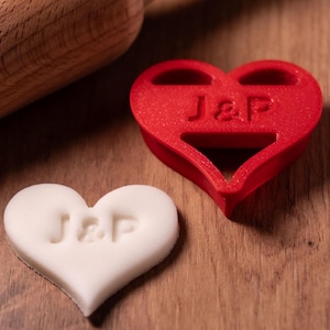 Könnte beinhalten: Ein roter herzförmiger Ausstecher mit den Initialen "J&P" eingraviert. Ein weißer herzförmiger Keks mit den gleichen Initialen ist ebenfalls abgebildet.