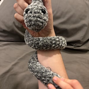 May include: A grey, plush crocheted snake with black button eyes. The snake is wrapped around an arm, showing its textured, soft material. The handmade toy is a unique, whimsical item.