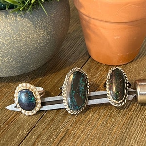 May include: Three silver rings with green and black opal stones set in a silver band. The rings are displayed on a wooden surface with a cactus and a potted plant in the background.