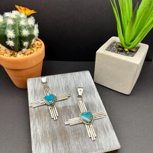 May include: Two silver pendants with turquoise heart-shaped stones in the center. The pendants are in the shape of a cross with radiating lines. The pendants are displayed on a wooden surface.
