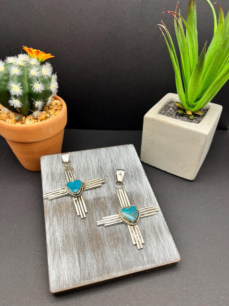 May include: Two silver pendants with turquoise heart-shaped stones in the center. The pendants are in the shape of a cross with radiating lines. The pendants are displayed on a wooden surface.