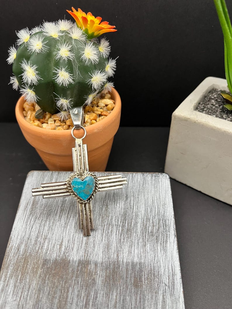 May include: A silver pendant with a turquoise heart in the center, surrounded by four silver bars. The pendant is in the shape of a cross.