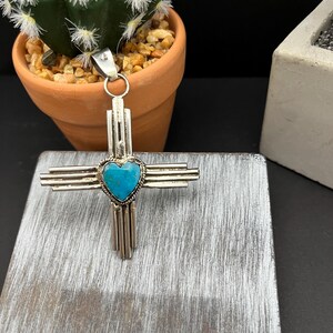 May include: A silver pendant in the shape of a cross with a turquoise heart in the center. The pendant is on a wooden surface.