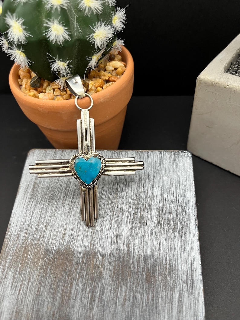May include: A silver pendant in the shape of a cross with a turquoise heart in the center. The pendant is on a wooden surface.