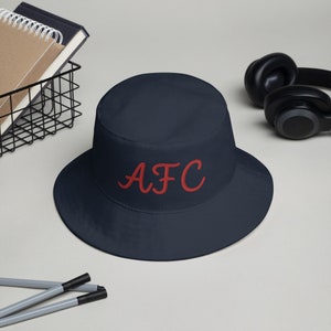 May include: A navy blue bucket hat with the word "AFC" embroidered in red on the front.