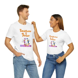 May include: Two white t-shirts with a cartoon image of a duck and a female duck dancing. The text on the shirts reads "Enseñame a Bailar".