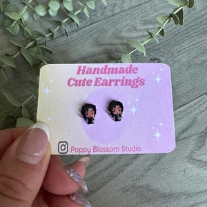 May include: A pair of handmade earrings featuring a cartoon character with black hair and a black outfit. The earrings are on a pink card with the text "Handmade Cute Earrings" and "Poppy Blossom Studio".