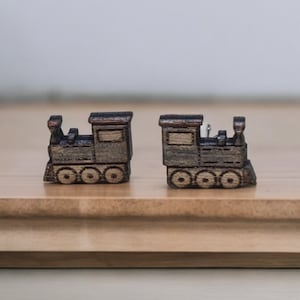 May include: Two miniature wooden train figurines. Each train is a detailed replica with a rectangular cabin, circular wheels, and a smokestack. The trains are a dark brown color and sit on a light brown wooden surface.