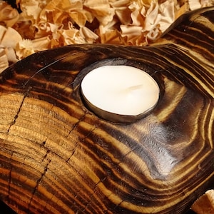 May include: A wooden candle holder with a dark brown and gold finish. The holder has a circular cutout for a tea light candle. The wood grain is visible and the holder has a rustic look.