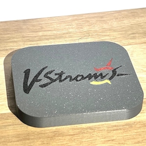 Pode incluir: Um descanso de copo quadrado cinza com o texto "V-Strom" em letras pretas e um design de estrela vermelha e amarela.